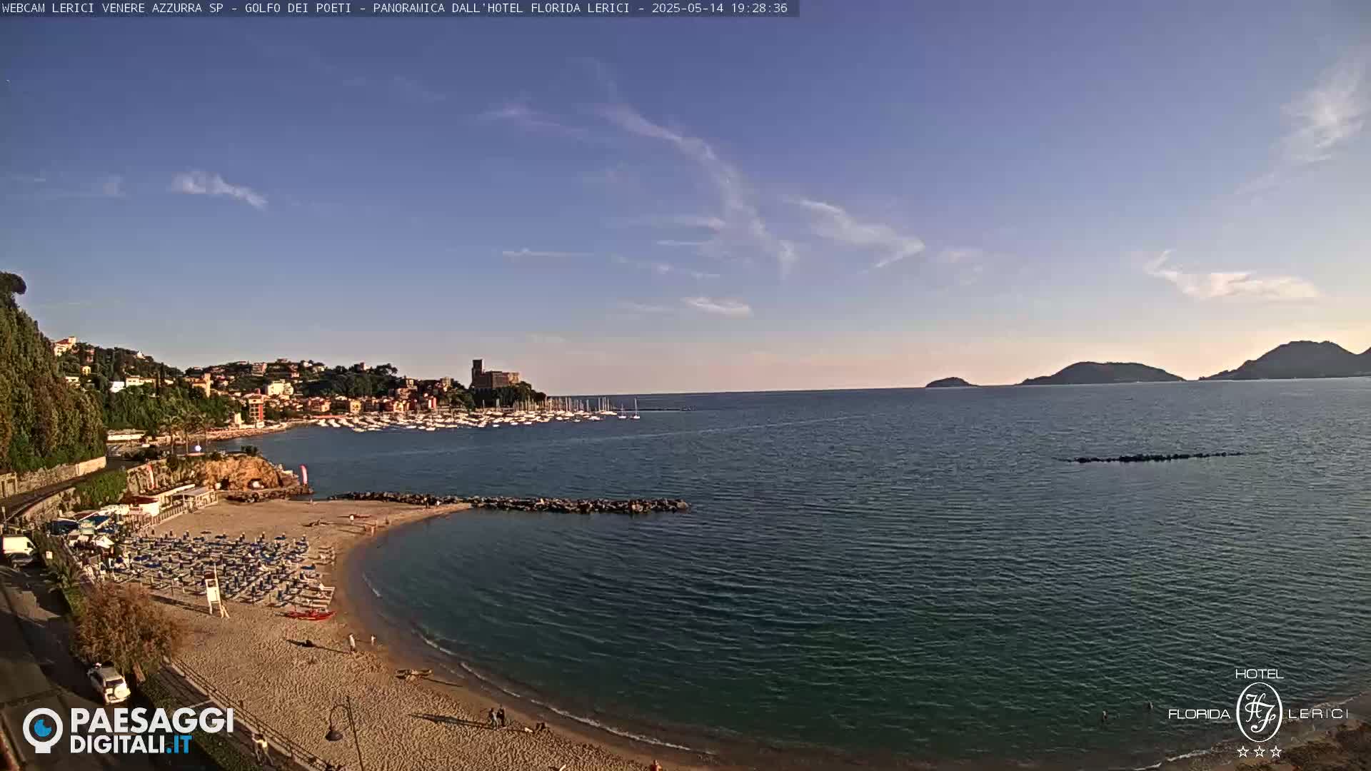 A sandy beach with rows of beach umbrellas and chairs is situated next to a calm body of water, with a town, harbor, and distant hills visible under a clear, mostly sunny sky.