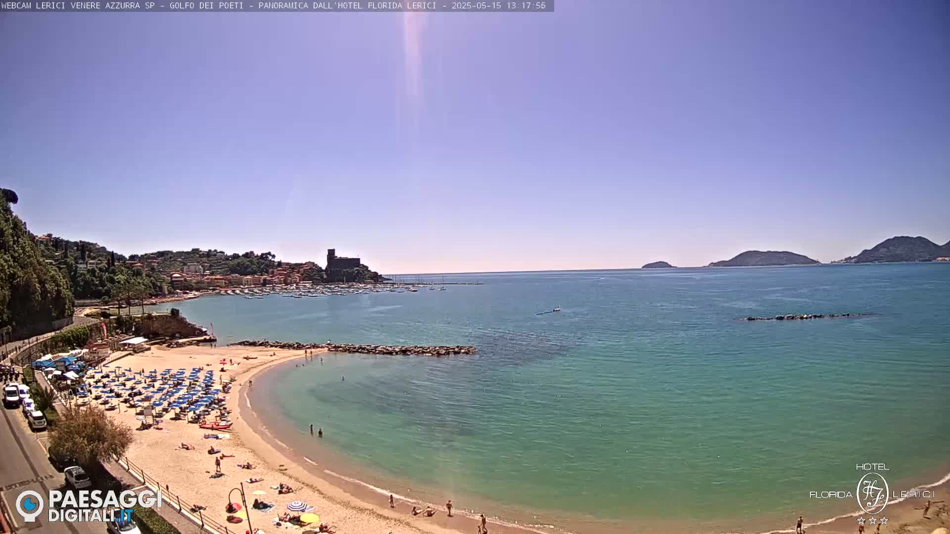 A sunny beach with numerous umbrellas and bathers curves into a calm, teal sea, backed by a town nestled against green hills and a distant island.