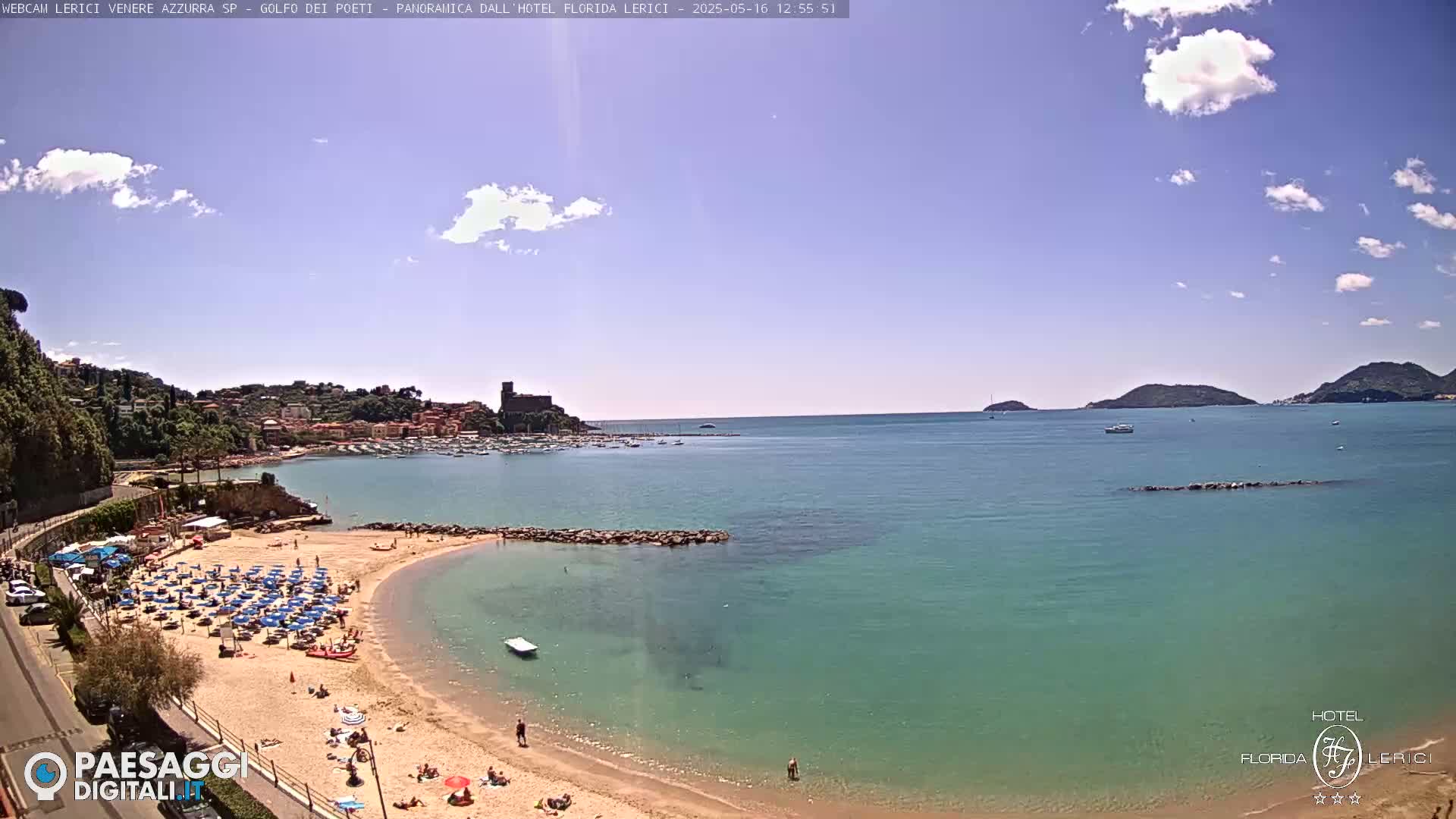 A sandy beach with blue umbrellas and sunbathers overlooks a calm, teal-colored sea under a mostly sunny sky.
