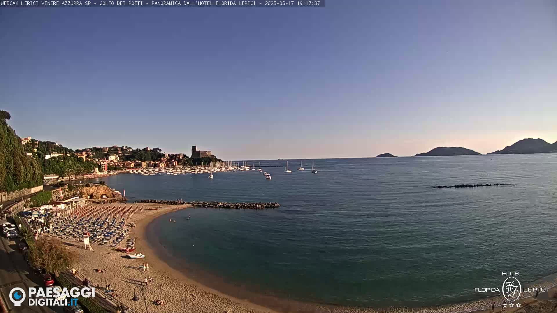 A sunny coastal scene shows a sandy beach with umbrellas and loungers, a calm bay with boats, and a town nestled against hills in the background.