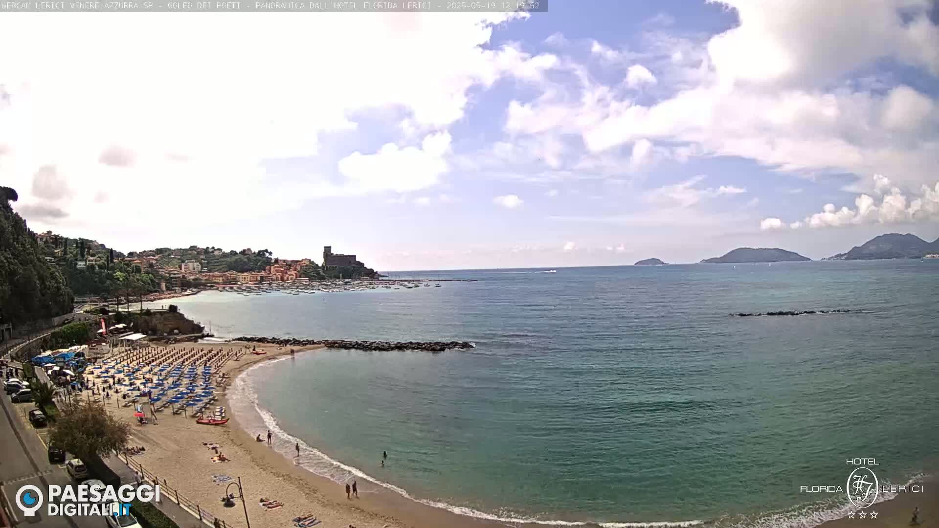 A partly cloudy day overlooks a sandy beach with many umbrellas and people, a calm bay with boats, and distant hills and islands.
