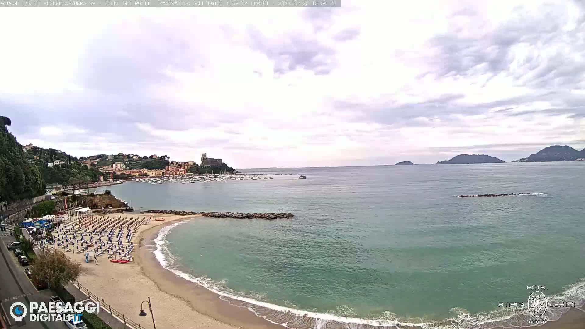 A mostly cloudy day shows a sandy beach with rows of closed umbrellas and chairs, a calm sea with a small breakwater, a town nestled on a hillside, and distant islands.
