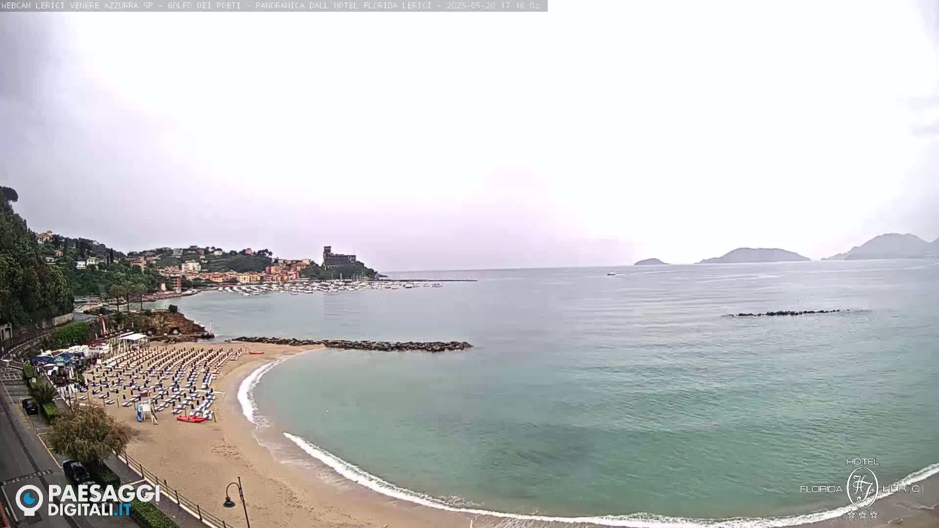 A beach with rows of lounge chairs is situated next to a calm, greenish-blue sea under an overcast sky, with a town and hills visible in the distance.