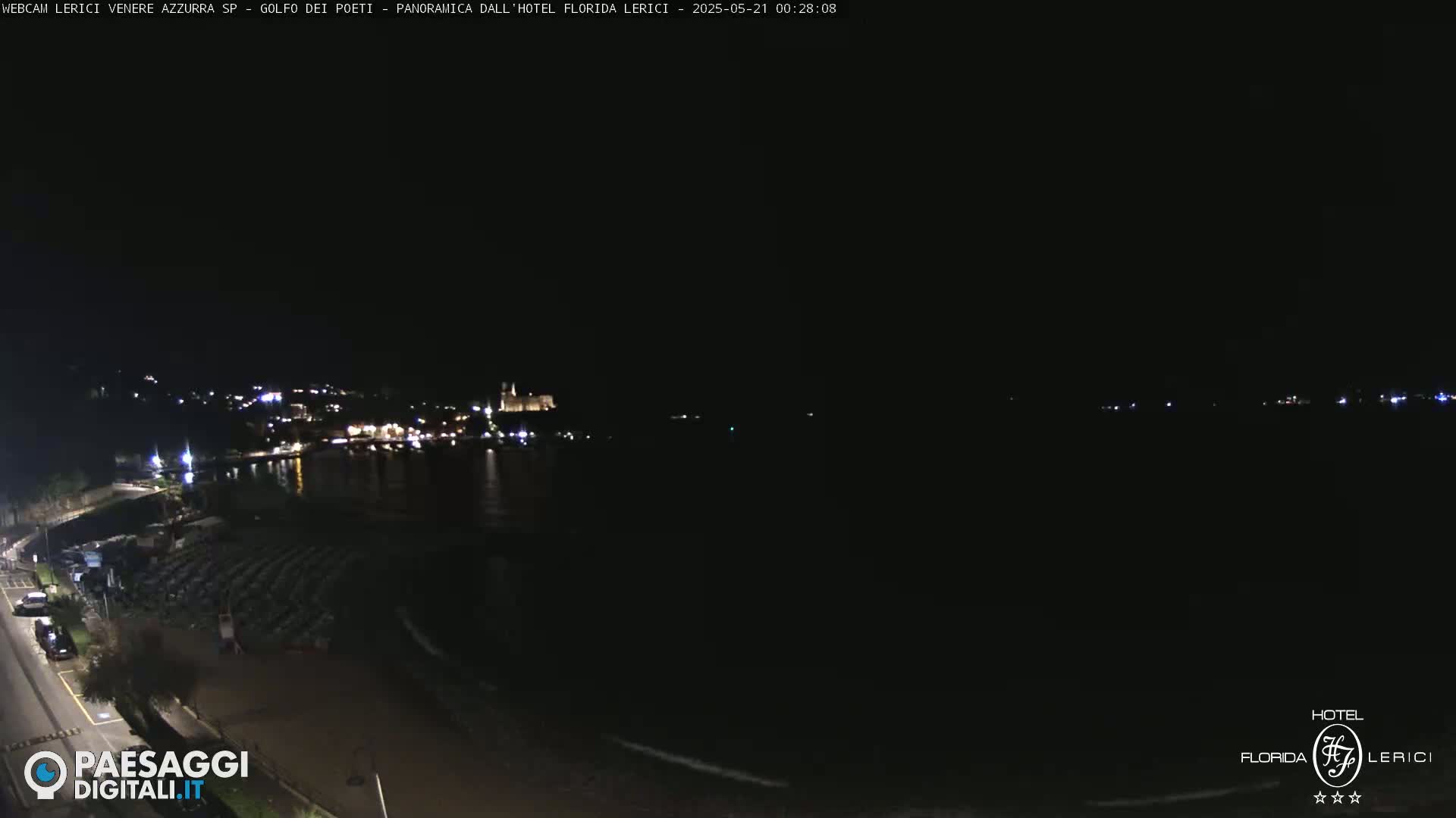 A nighttime coastal scene shows a dark body of water with a dimly lit town across the water, and a dark beach with rows of folded chairs in the foreground.