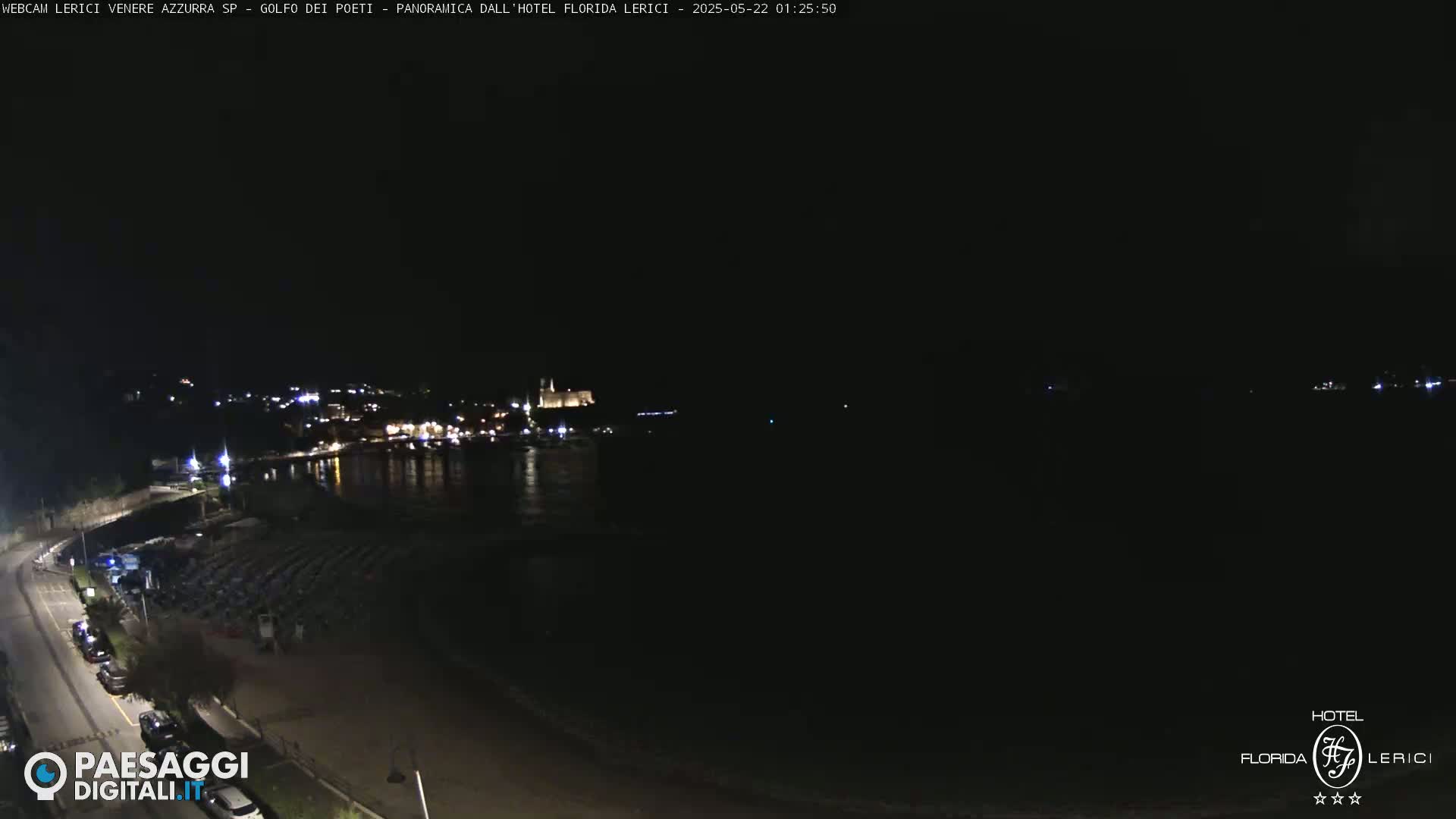 A nighttime, seaside view shows a dark beach with folded beach chairs, a road with parked cars, and a dimly lit town across the water.