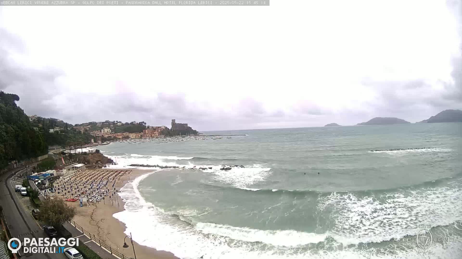 A sandy beach with rows of beach umbrellas is partially visible beneath an overcast sky, with moderate waves rolling into the shore of a bay and some distant hills.
