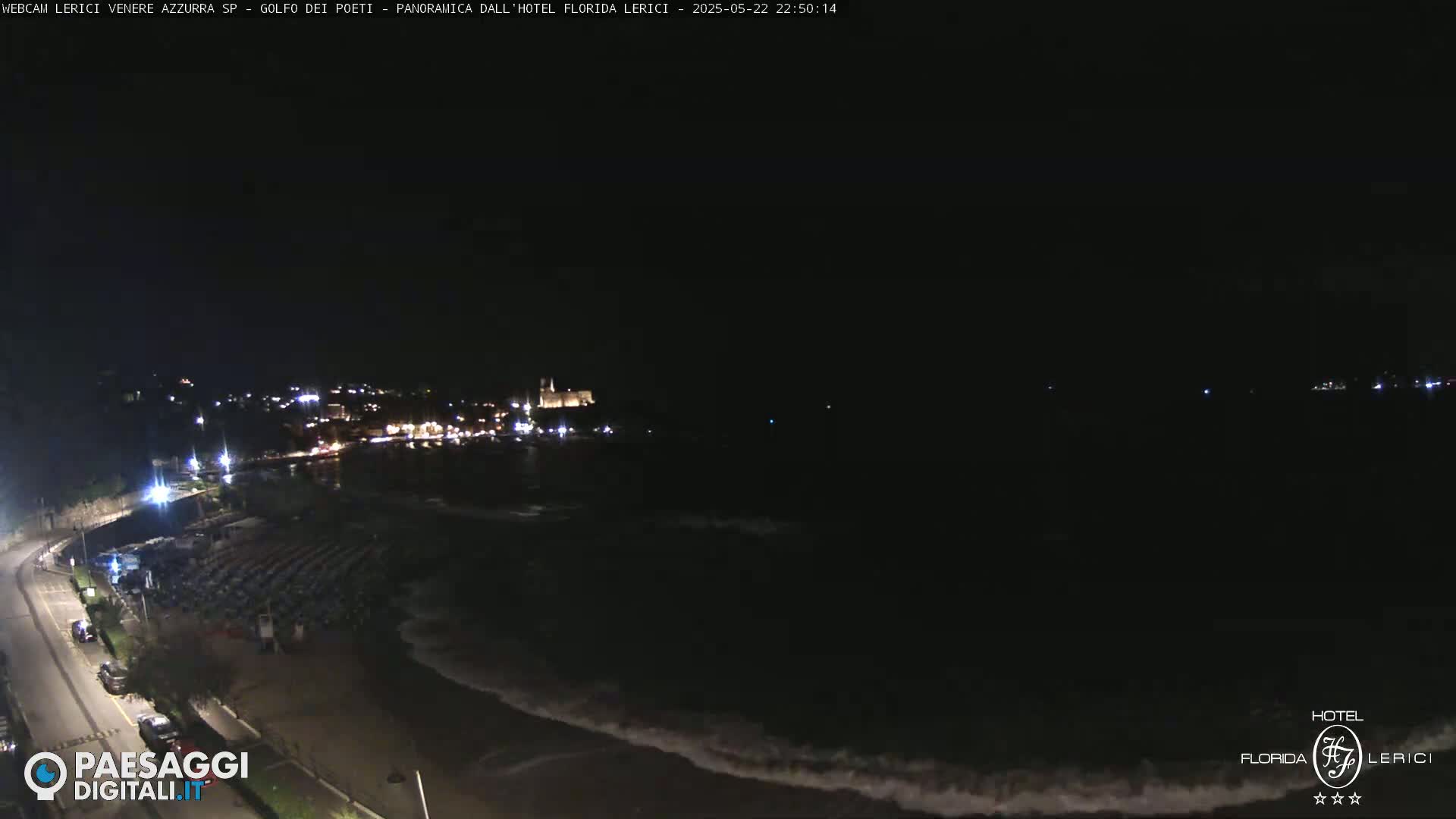 A nighttime coastal scene shows a dark ocean with waves lapping the shore, a road with parked cars, and dimly lit buildings in the distance.