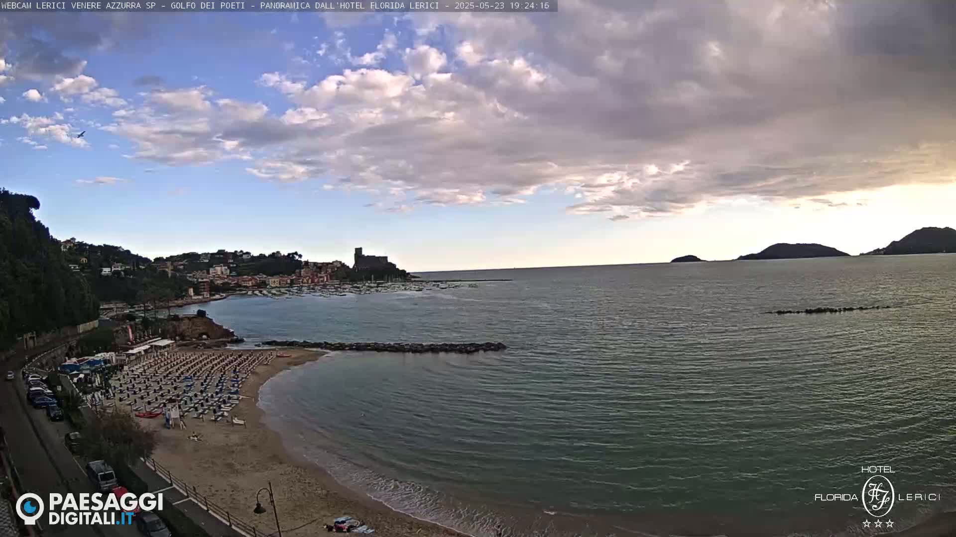 A partly cloudy sky overlooks a sandy beach with numerous umbrellas and chairs, a calm sea, and a town nestled on a hillside in the distance.