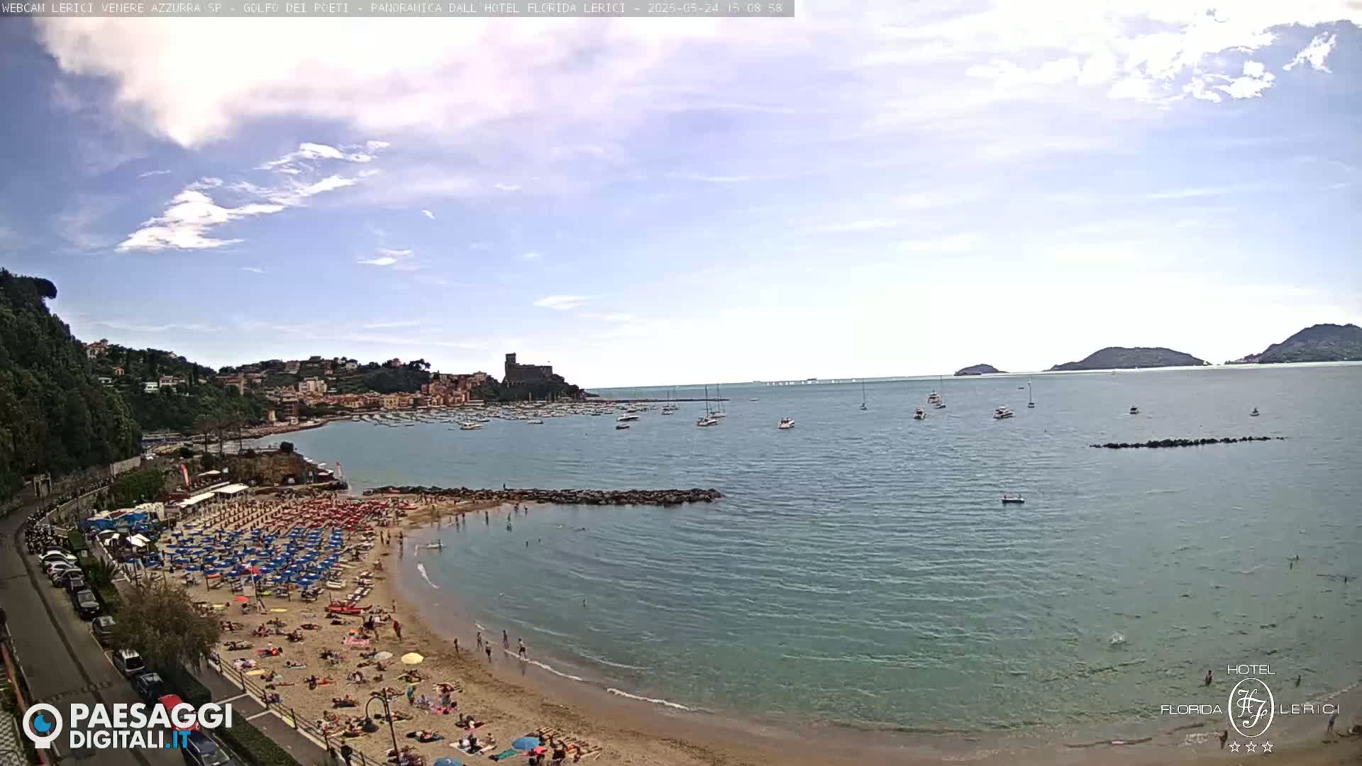 A sunny day shows a crowded beach with blue umbrellas and calm, blue-green water with boats and islands in the distance.