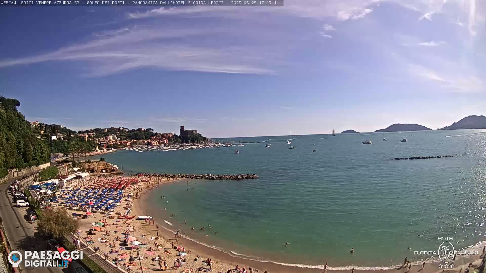 A sunny day at a beach shows a sandy shore crowded with people, umbrellas, and calm turquoise water, with boats and hills in the background.