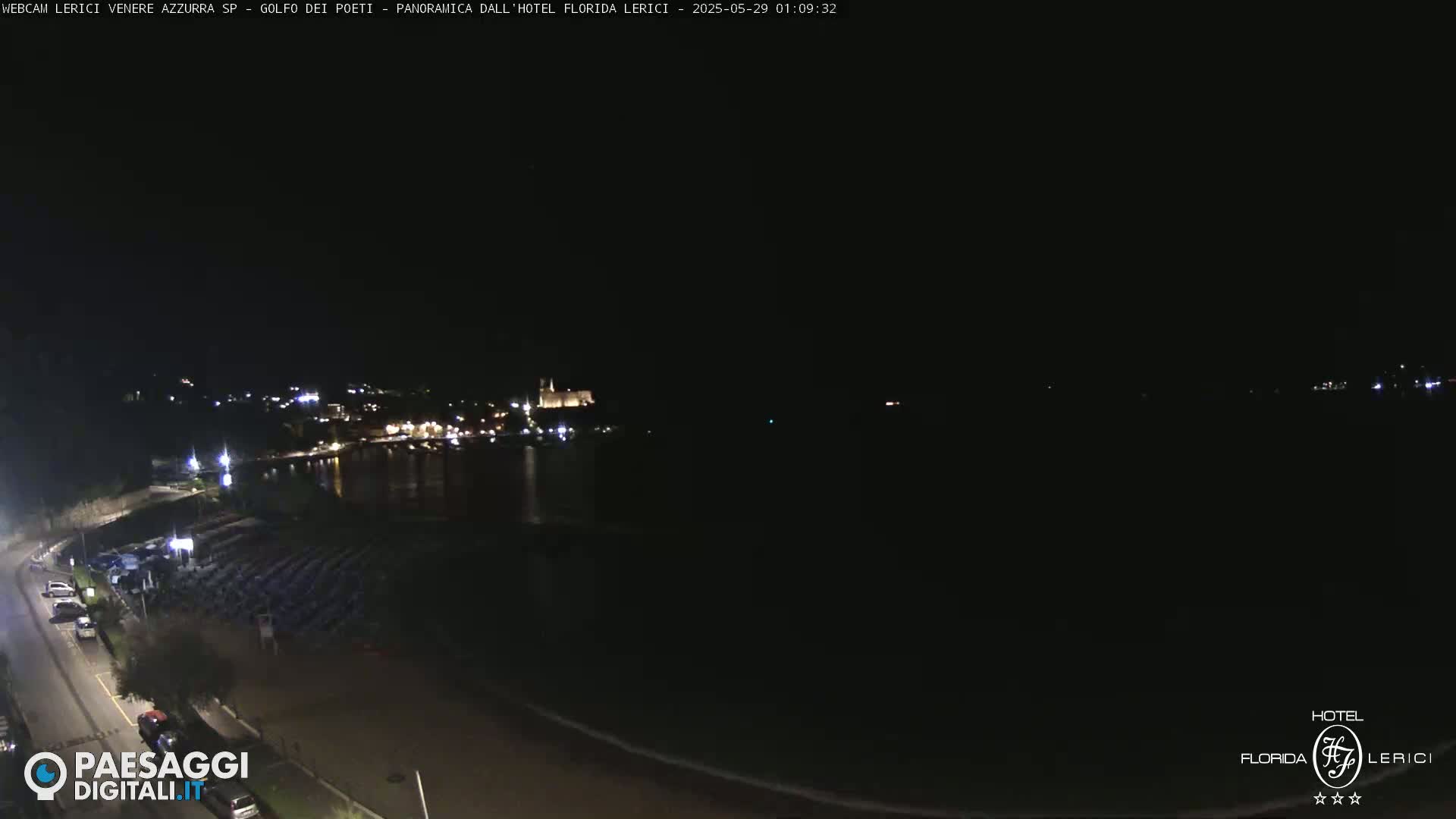 A nighttime, high-angle view shows a dark body of water with a town's lights faintly visible in the distance, a beach with rows of dark beach chairs, and a road with a few cars parked along the side.
