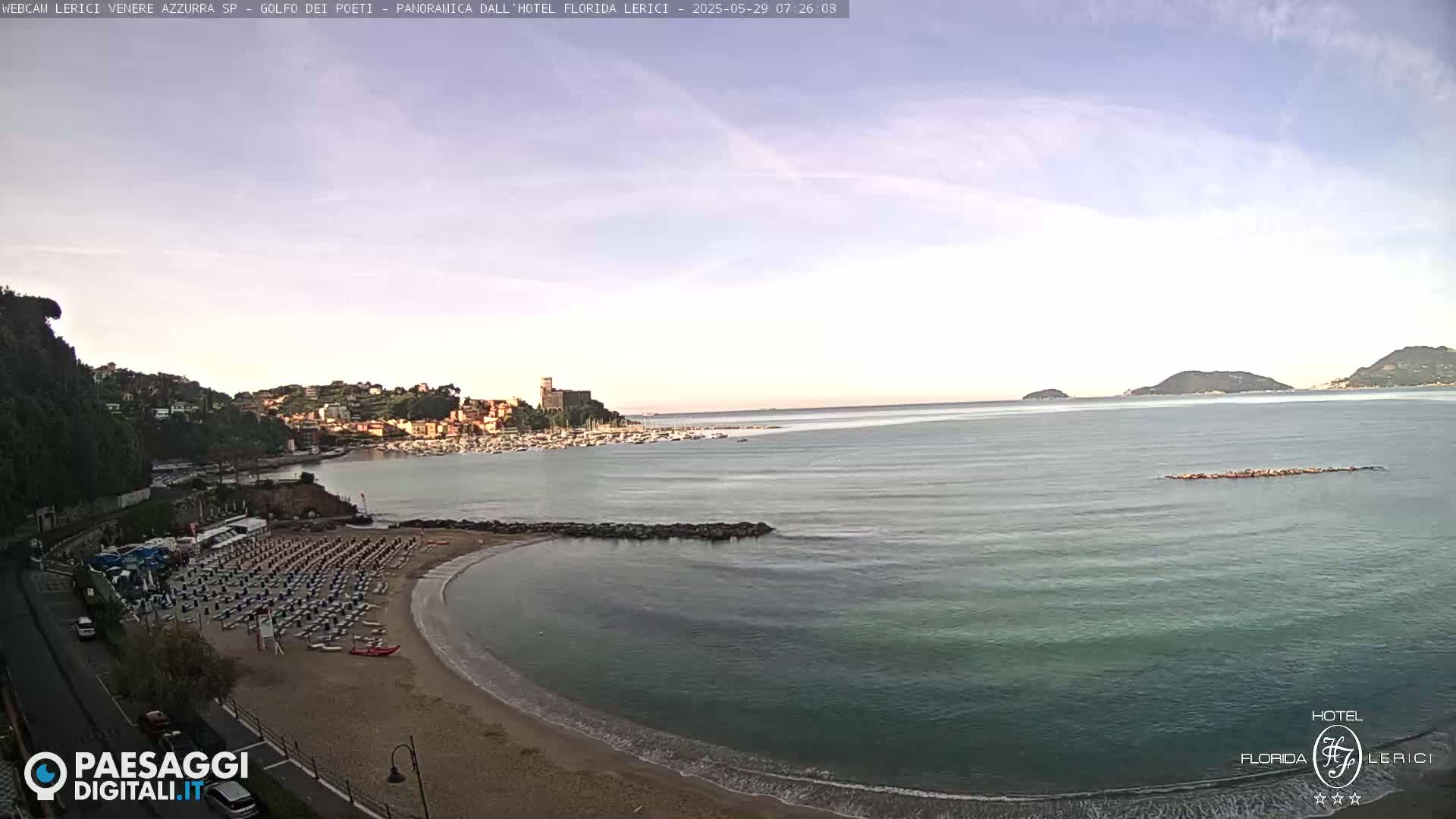 A mostly sunny day shows a sandy beach with rows of beach chairs next to calm, teal water and a small town across the bay under a pale sky.