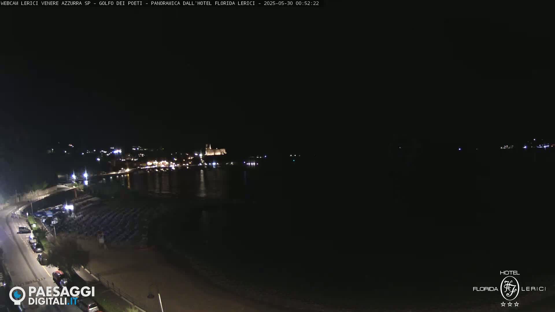A nighttime coastal scene shows a dark body of water reflecting distant lights from a town, with a beach and parked cars visible in the foreground under a clear, dark sky.