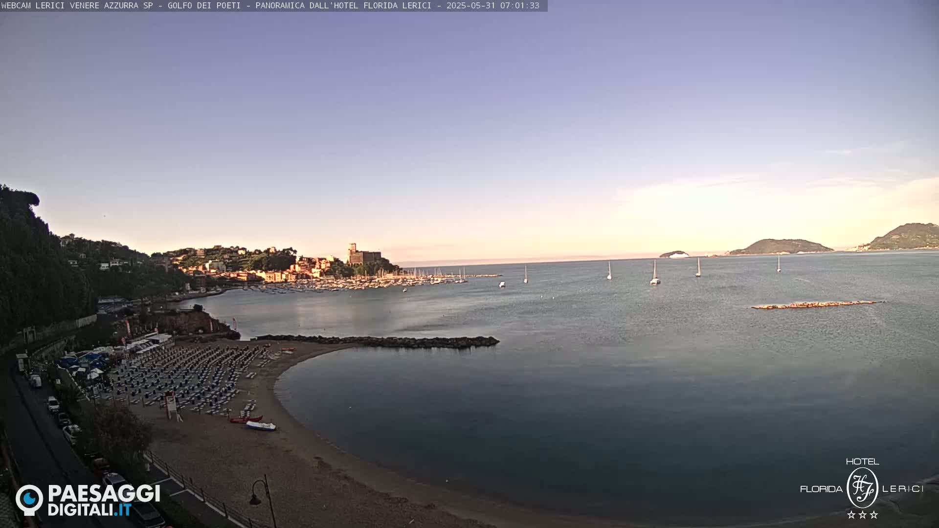 A calm, clear morning shows a sandy beach with rows of empty lounge chairs, a calm bay with moored boats, and a town nestled on a hillside beyond.