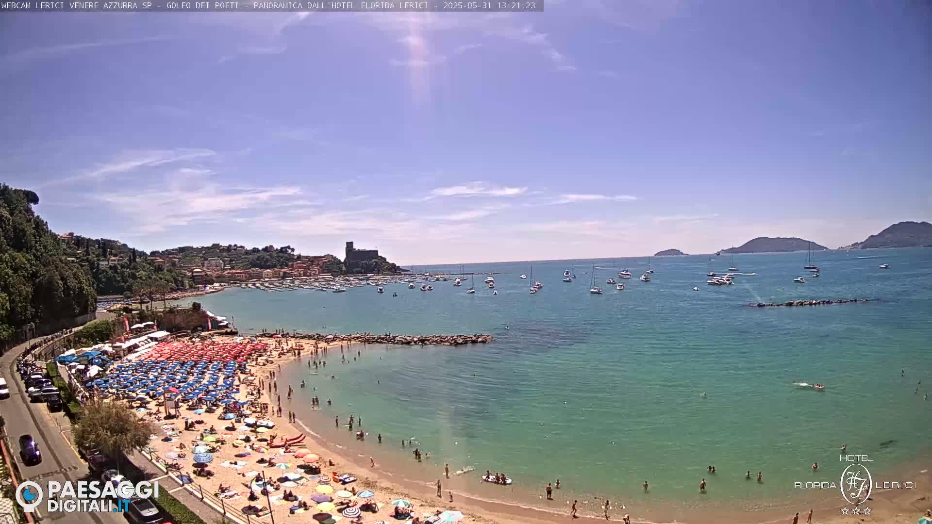 A sunny day at a crowded beach with numerous umbrellas, people swimming and sunbathing, boats in the bay, and a town nestled on a hillside in the background.