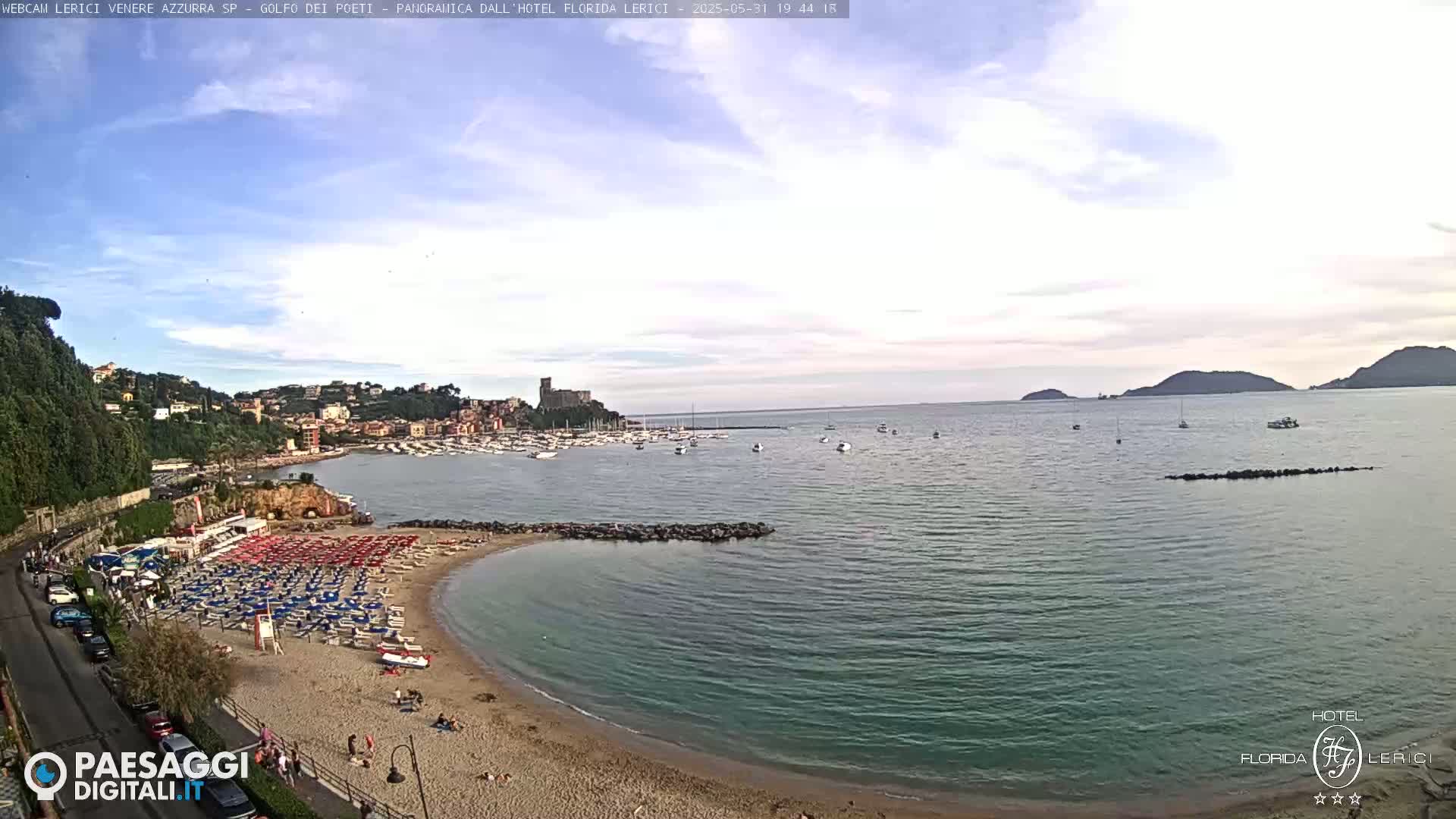 A partly cloudy day shows a sandy beach with colorful umbrellas and chairs next to a calm, teal-colored bay, with a town and boats visible in the distance.