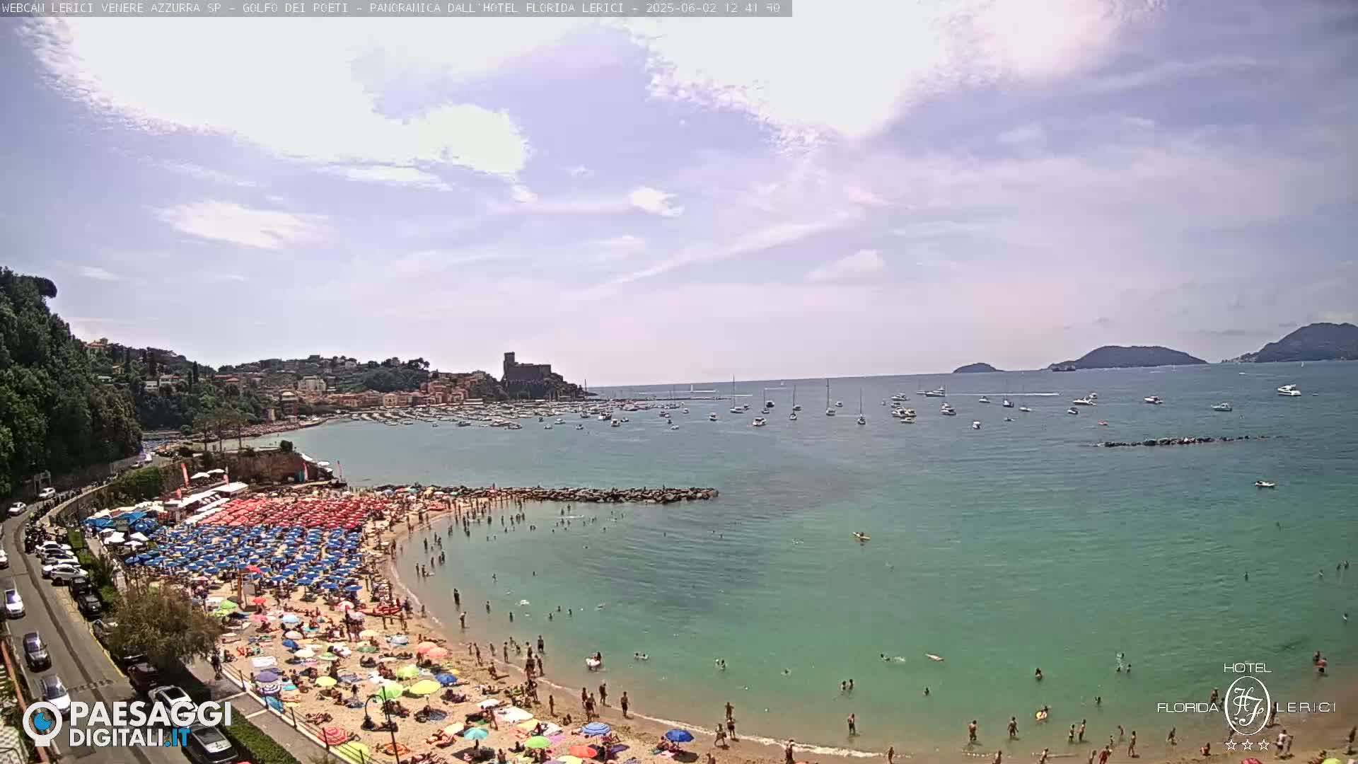 A crowded beach with colorful umbrellas and turquoise water under partly cloudy skies, boats are visible in the bay beyond a rocky breakwater.