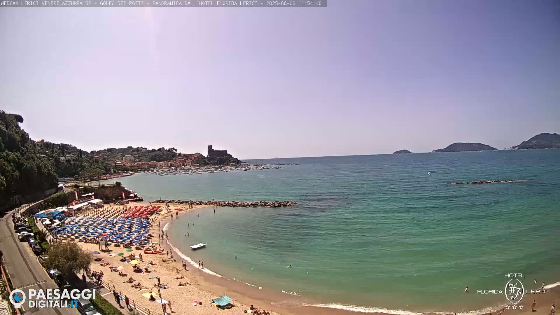 A sunny day at a beach, showing a sandy shore lined with colorful umbrellas and sunbathers, a calm teal sea, and a town nestled on a hillside in the distance.