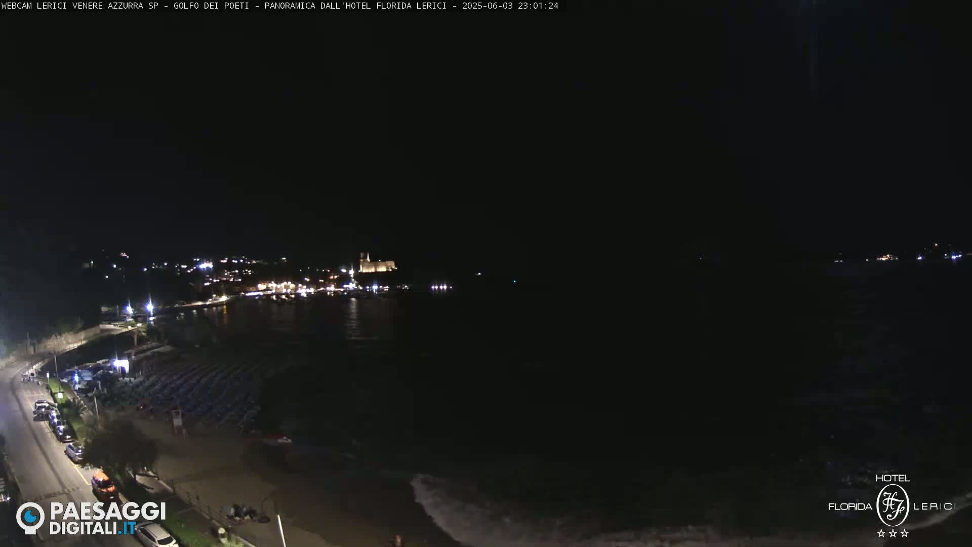 A nighttime coastal scene shows a dark body of water with a town's lights visible in the distance, along with a beach with rows of folded-up beach chairs and a road with cars parked.