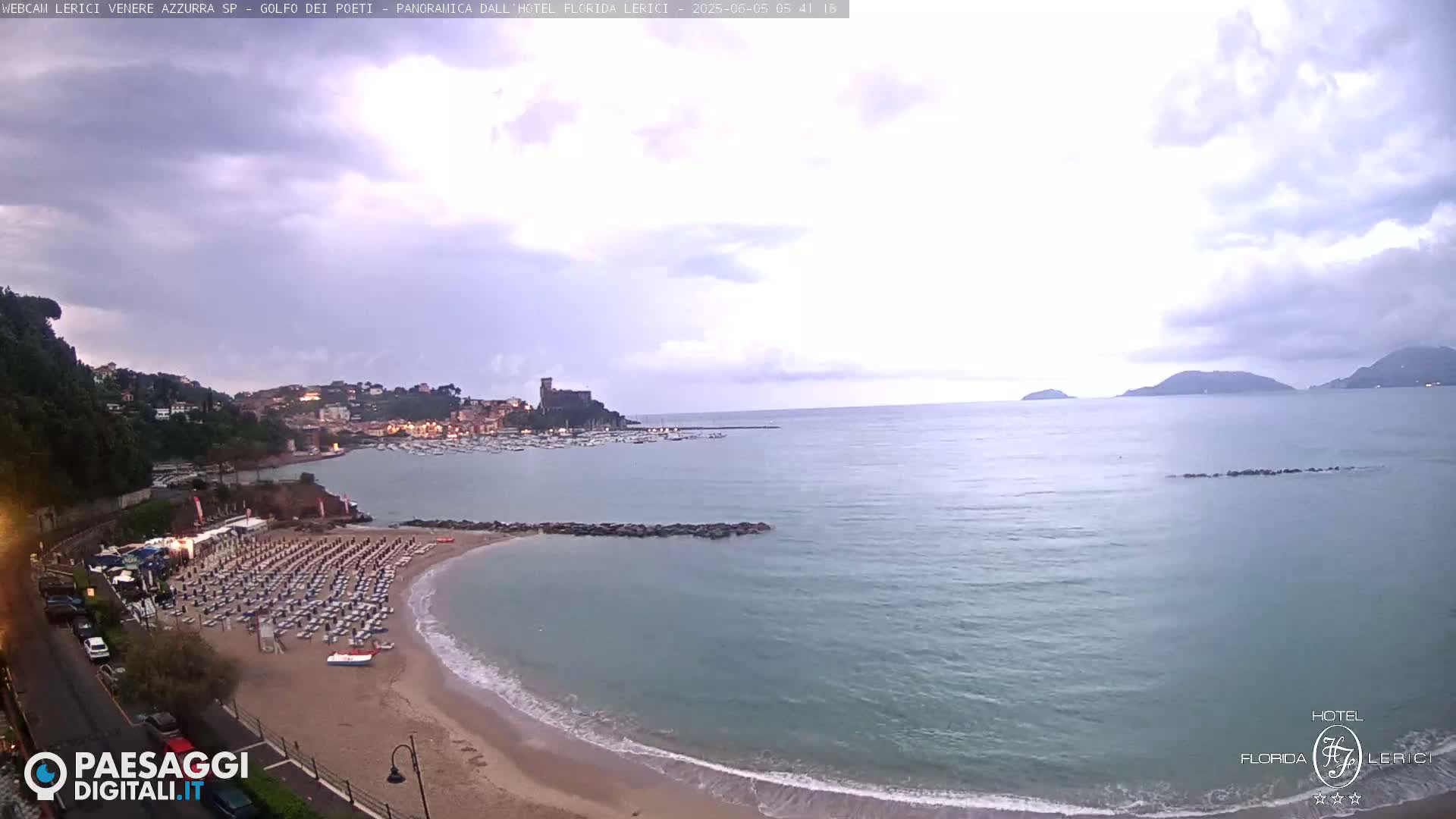 A mostly cloudy day reveals a sandy beach with rows of lounge chairs, a calm sea with a small harbor full of boats, and a town nestled along a hillside.