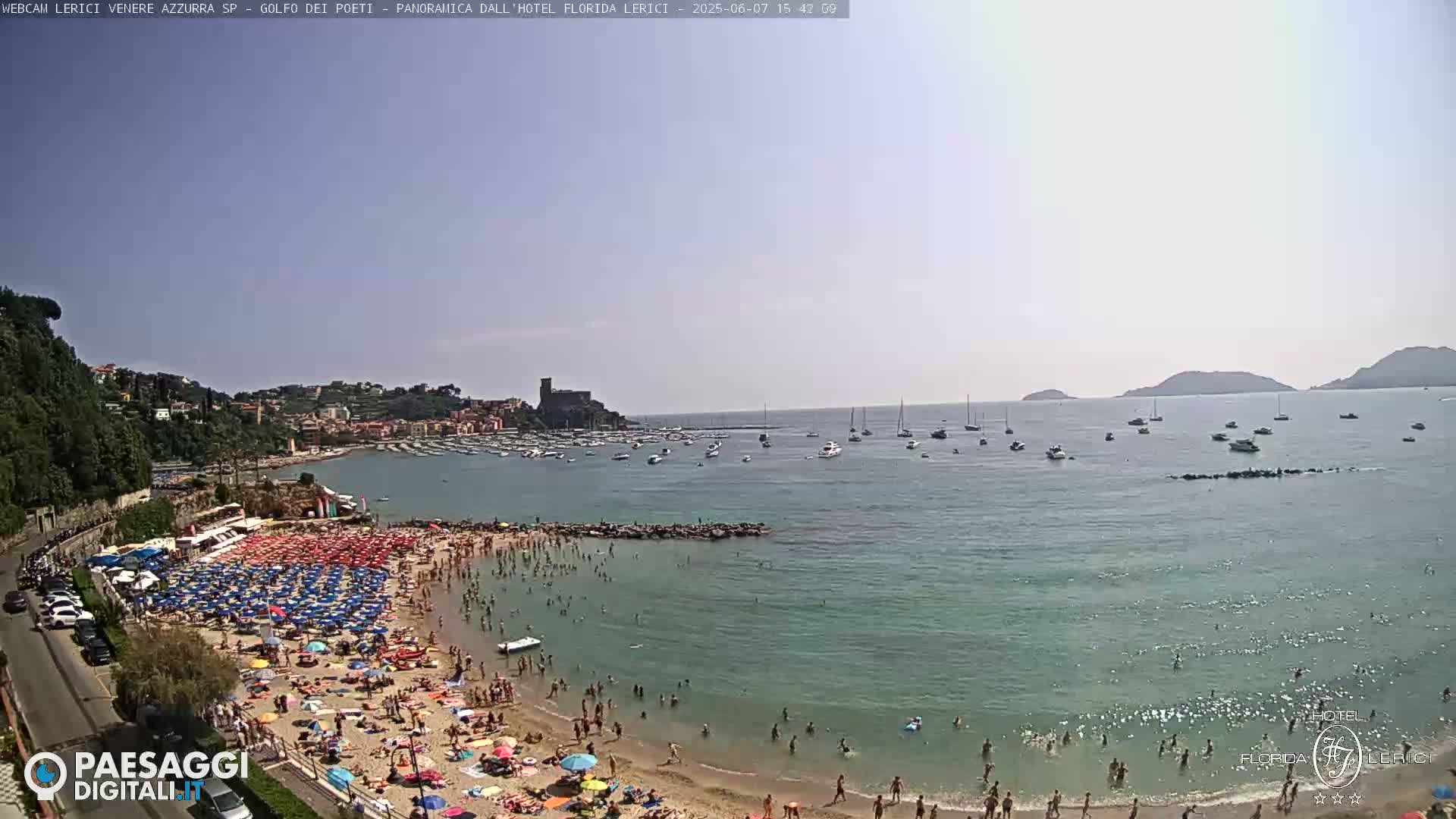 A crowded beach with numerous colorful umbrellas and people swimming in calm, clear water under a mostly clear sky, boats are visible in the bay beyond the beach.