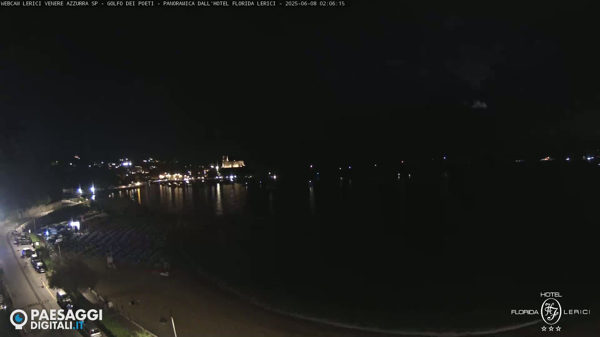 A nighttime view of a dark beach and calm water, with a town's lights faintly visible in the distance.