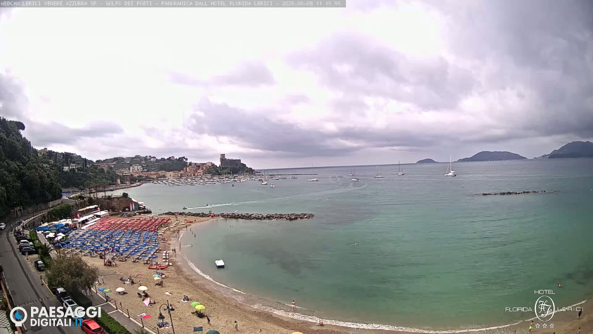 A sandy beach with numerous red and blue umbrellas, a calm teal sea with boats, and a cloudy sky overlooks a town nestled against hills.