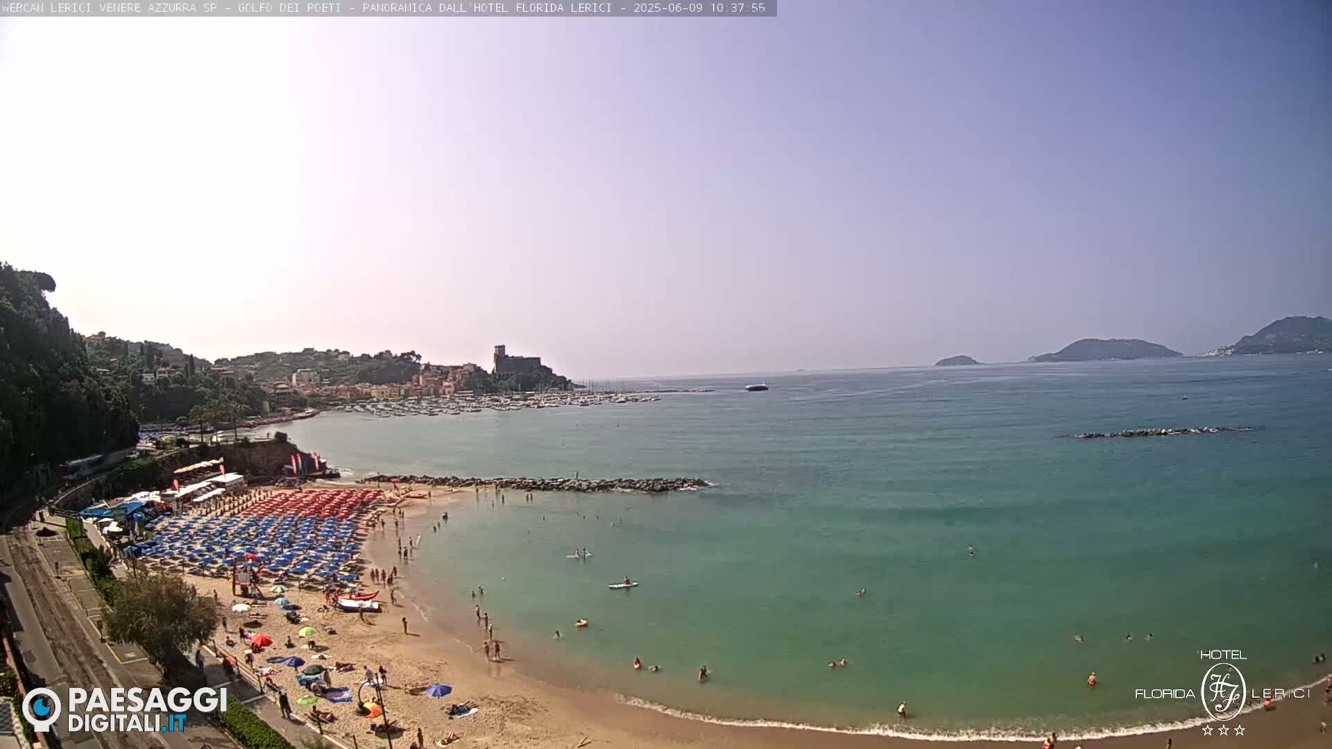 A sunny day at a sandy beach with clear turquoise water, numerous umbrellas, and people swimming and sunbathing, overlooking a town and a calm bay.