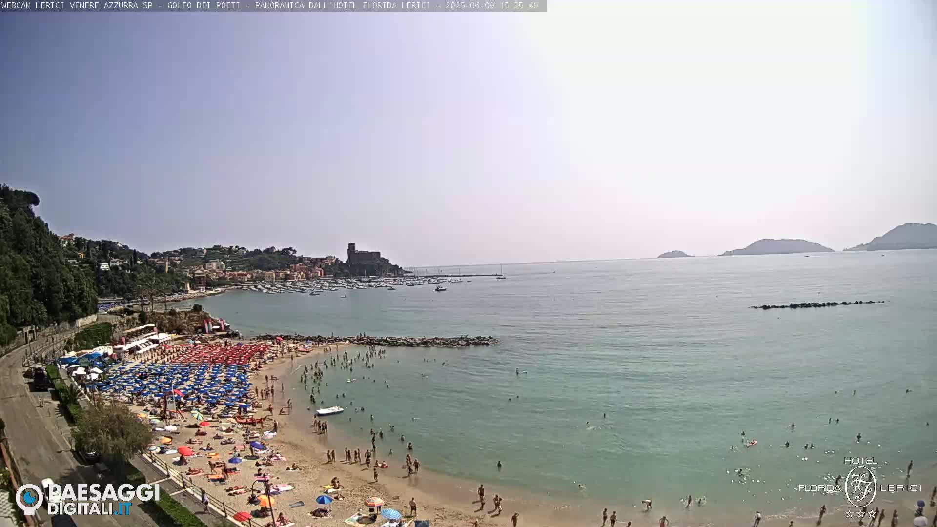 A sunny day shows a crowded beach with numerous umbrellas and people swimming in a calm sea, overlooking a town with boats in the harbor and distant hills.