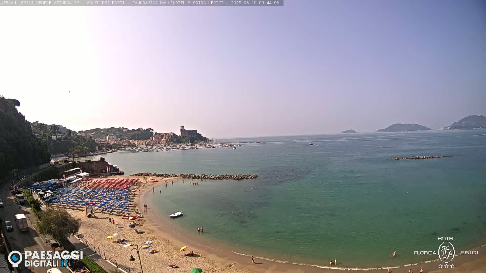 A sunny coastal scene shows a sandy beach with numerous colorful umbrellas and people enjoying the water, a town nestled against a hillside, and calm, clear water extending to distant islands.