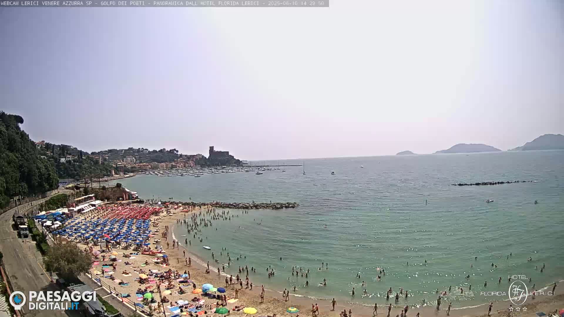 A crowded beach with numerous umbrellas and people swimming in calm, clear water under a hazy sky, backed by a town and hills.