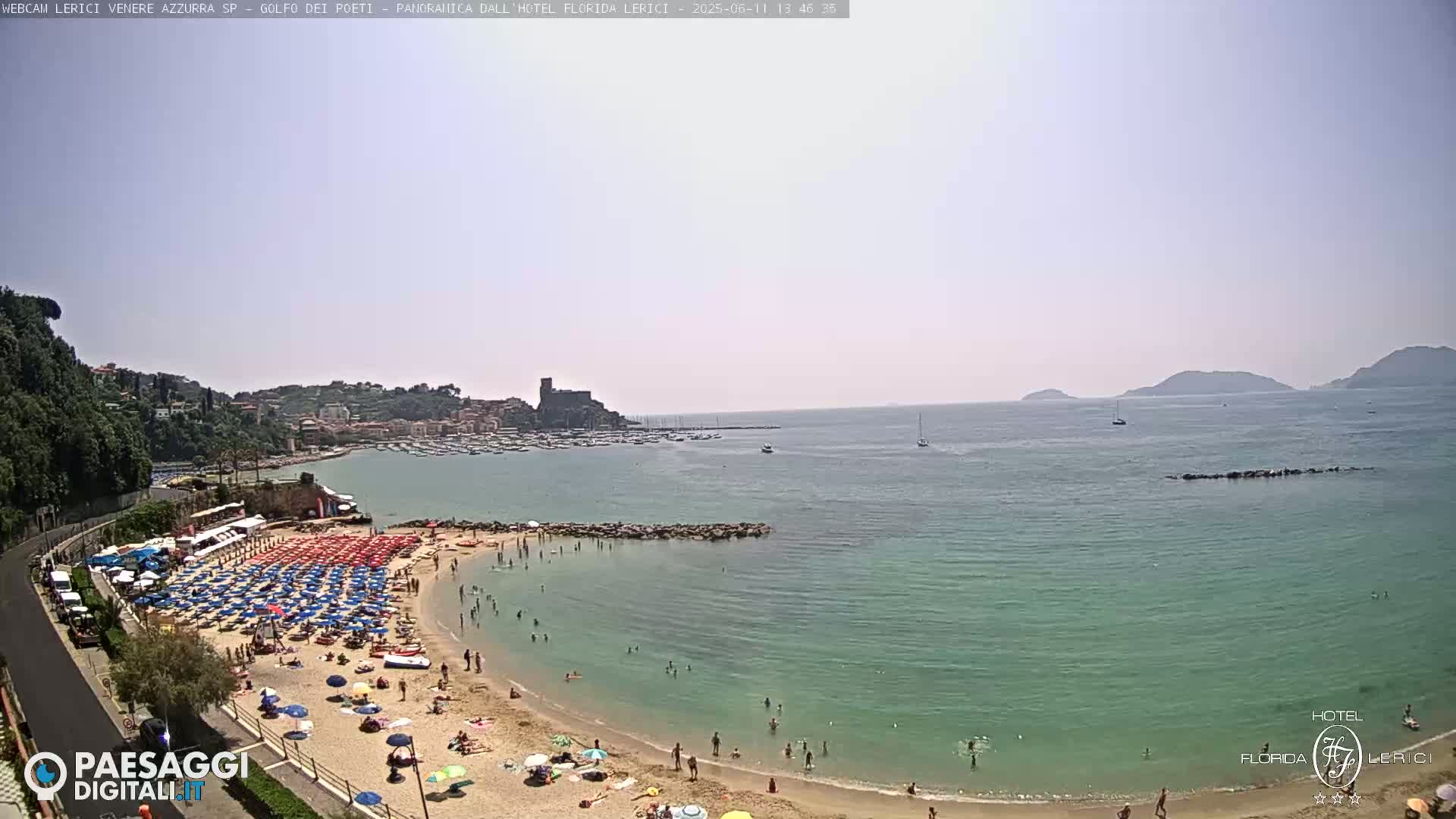 A sunny day at a beach with a calm, teal-colored sea, a sandy shore lined with red and blue beach umbrellas and bathers, and a town visible in the distance nestled against green hills.