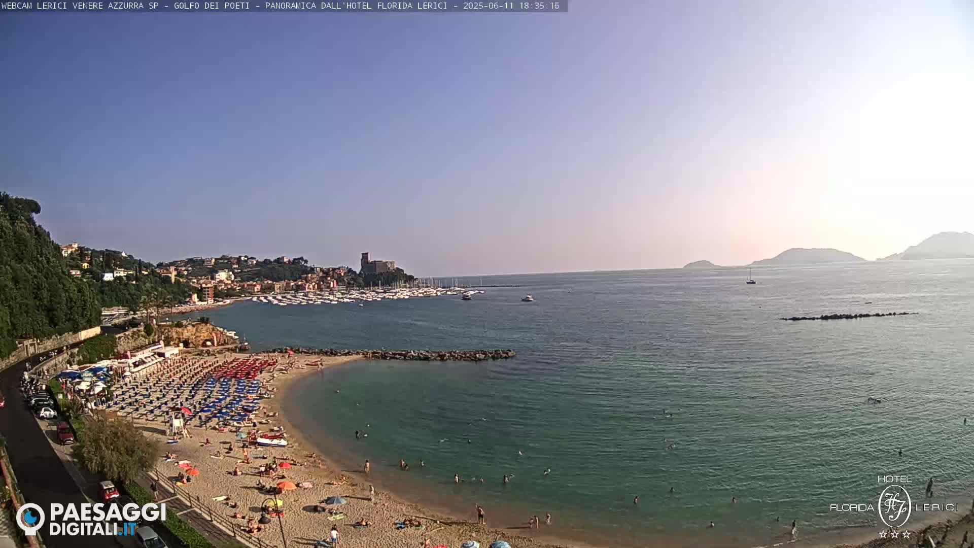 A sunny day at a beach with calm, clear water, showing a coastline with buildings and boats, and numerous people relaxing on the sand.