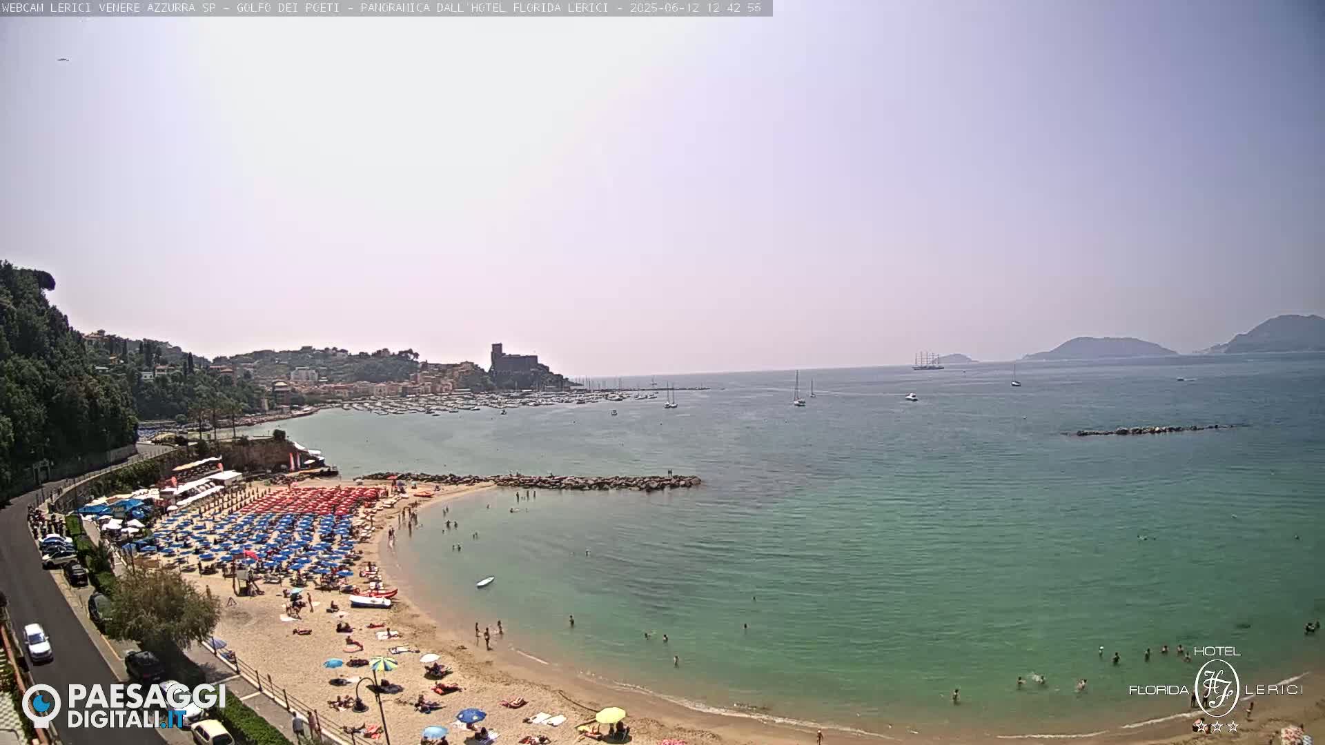 A sunny beach with numerous umbrellas and people is situated next to calm, teal water, backed by a town and distant hills.