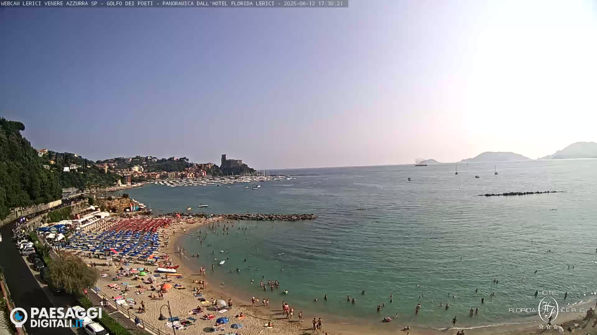 A sunny day at a crowded beach with clear water, boats in the bay, and a town nestled on a hillside in the background.