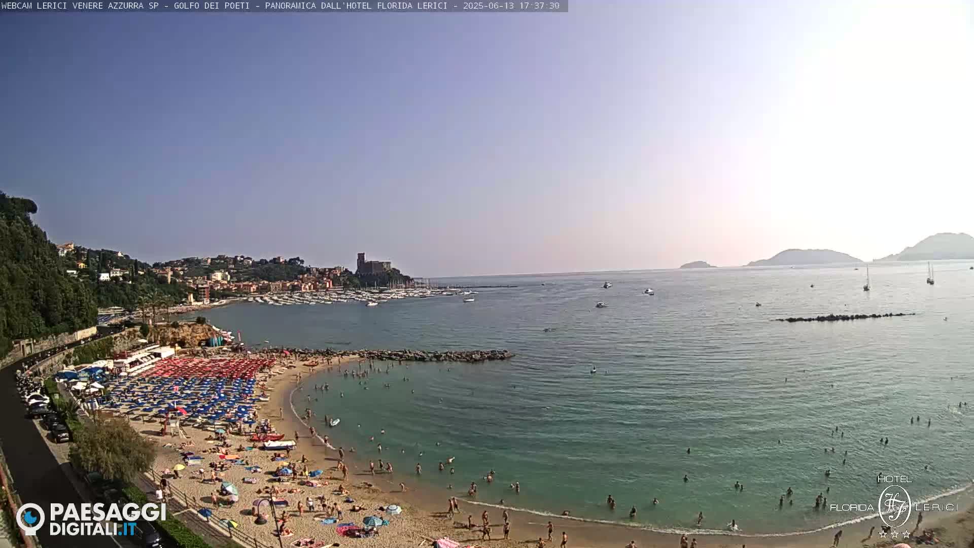 A sunny day at a crowded beach with calm, clear water, featuring numerous colorful umbrellas and people swimming and sunbathing.