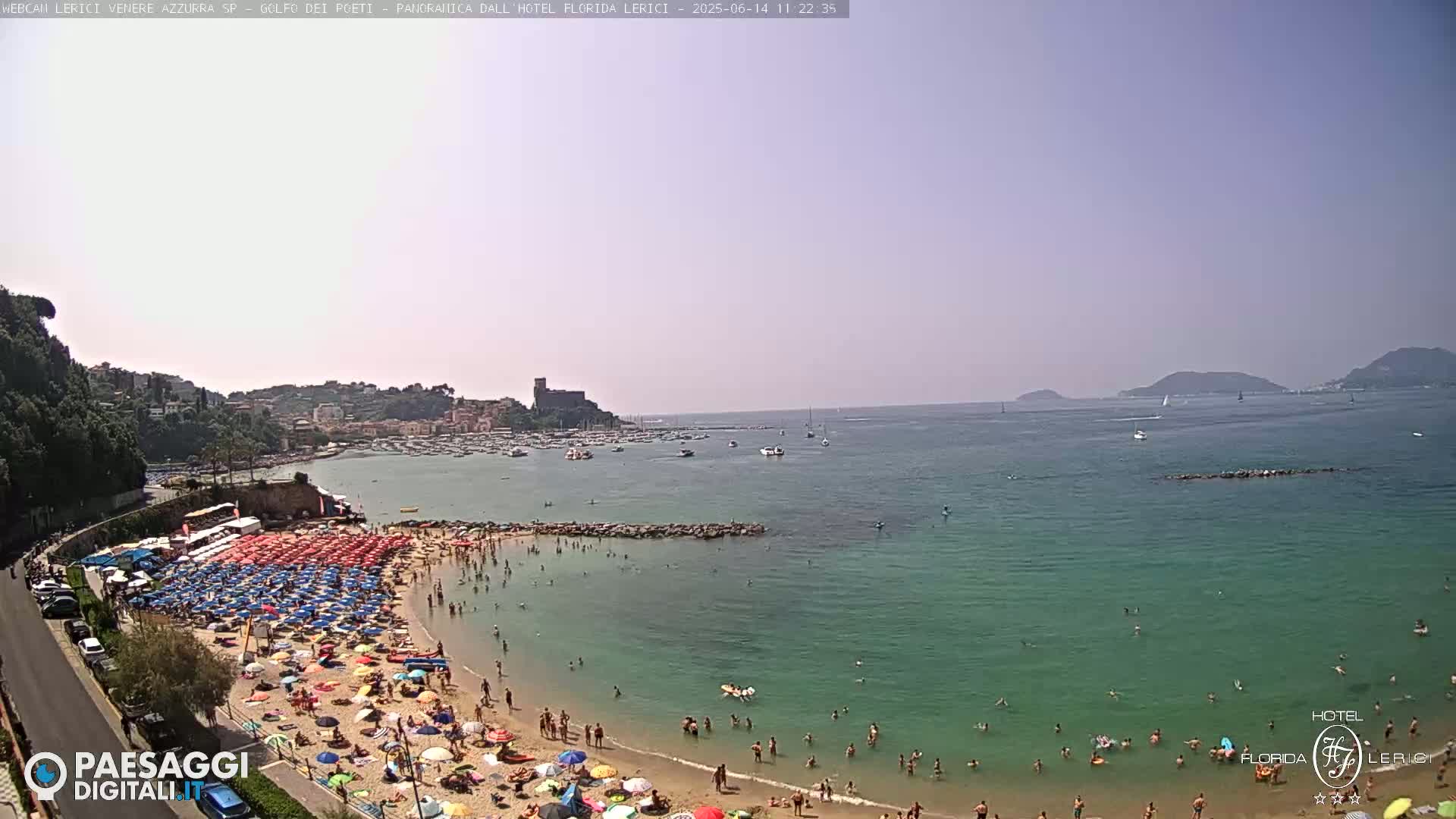 A sunny day at a crowded beach with turquoise water, numerous colorful umbrellas, and a town with a castle visible in the distance.