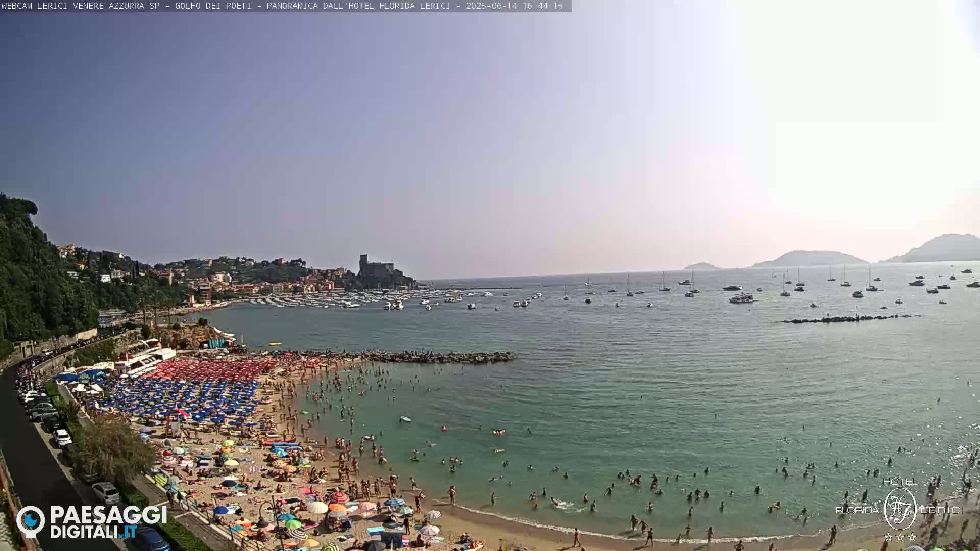 A crowded beach with numerous colorful umbrellas under a sunny sky overlooks a calm bay filled with boats and distant hills.