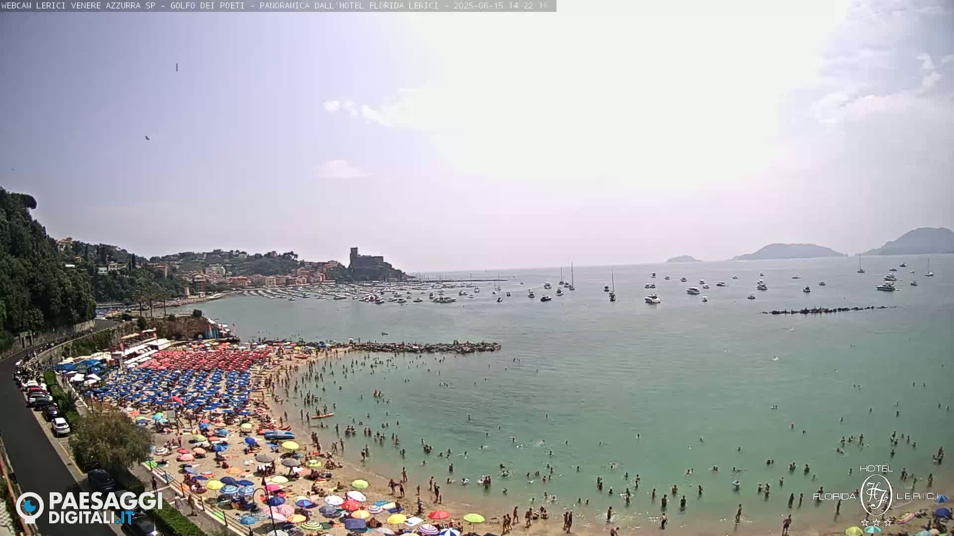 A crowded beach with numerous colorful umbrellas under a mostly clear sky, adjacent to a calm bay filled with boats and a distant town.