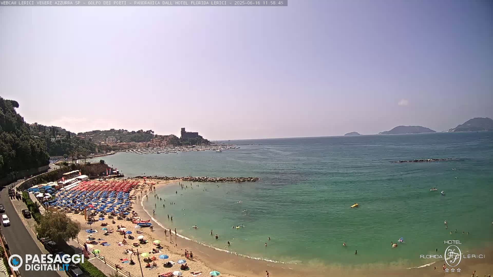 A sunny day at a crowded beach with turquoise water, numerous colorful umbrellas, and a town nestled on a hillside in the background.
