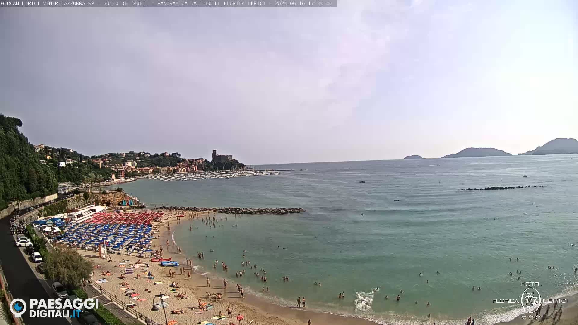 A sandy beach with numerous umbrellas and sunbathers is situated next to calm, clear ocean water under a mostly clear sky.