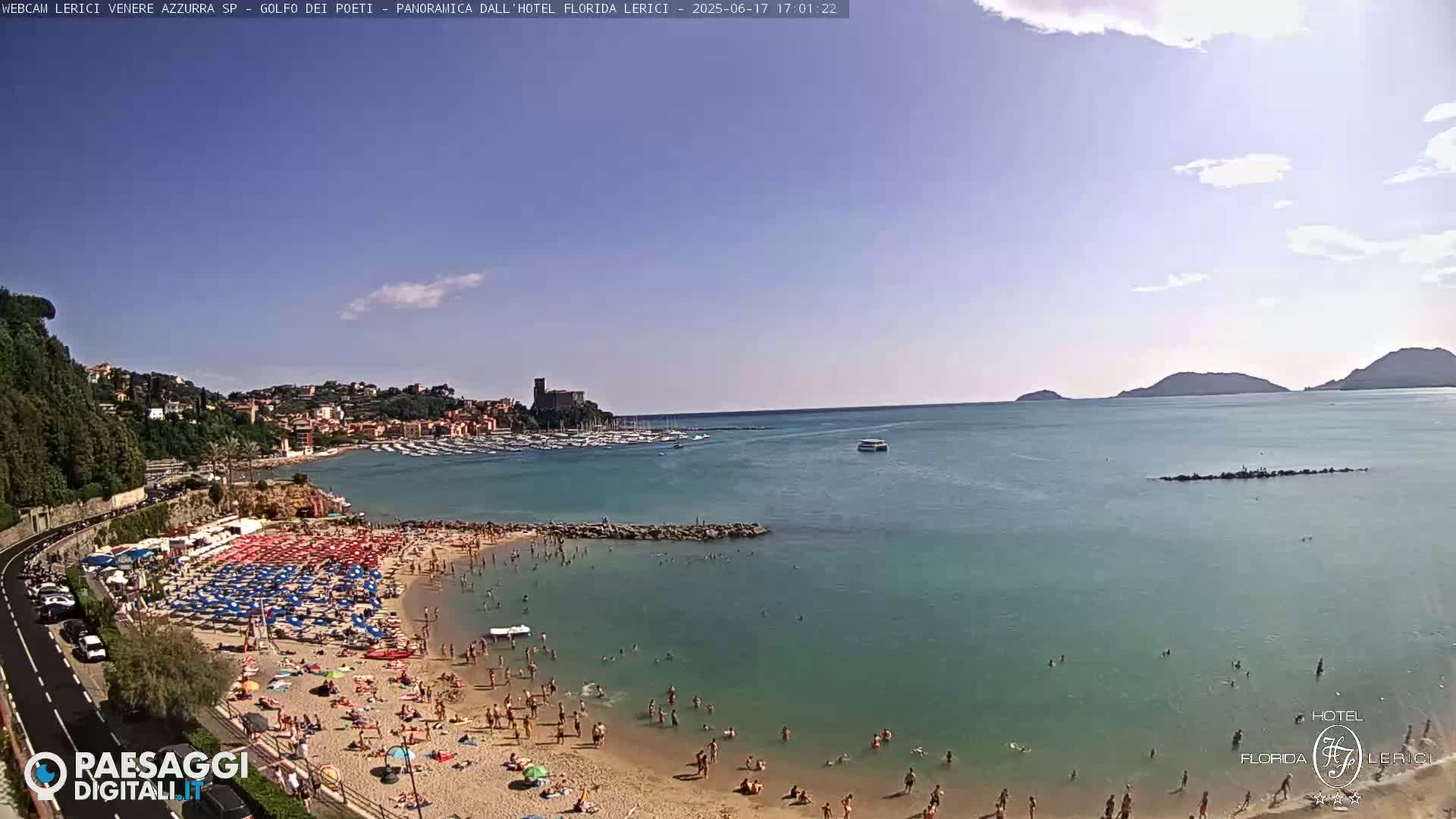 A crowded beach with colorful umbrellas and people swimming in clear, shallow water next to a calm bay, under a mostly sunny sky.