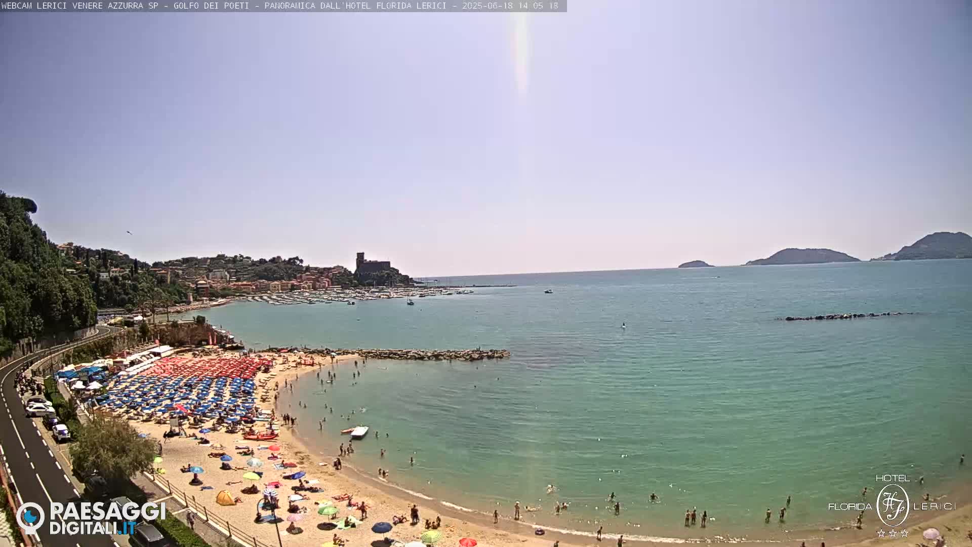 A sunny day at a crowded beach with calm, teal water, boats in a harbor, and a town nestled on a hillside in the background.