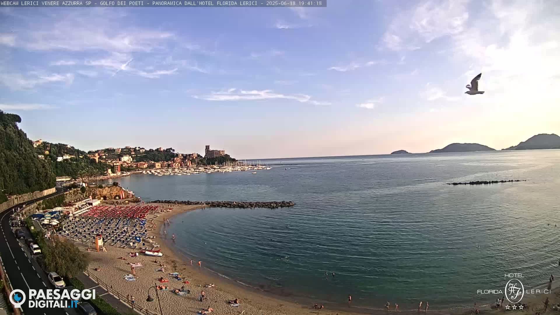 A partly cloudy sky overlooks a calm bay with a sandy beach filled with people and umbrellas, a town in the background, and a seagull in flight.
