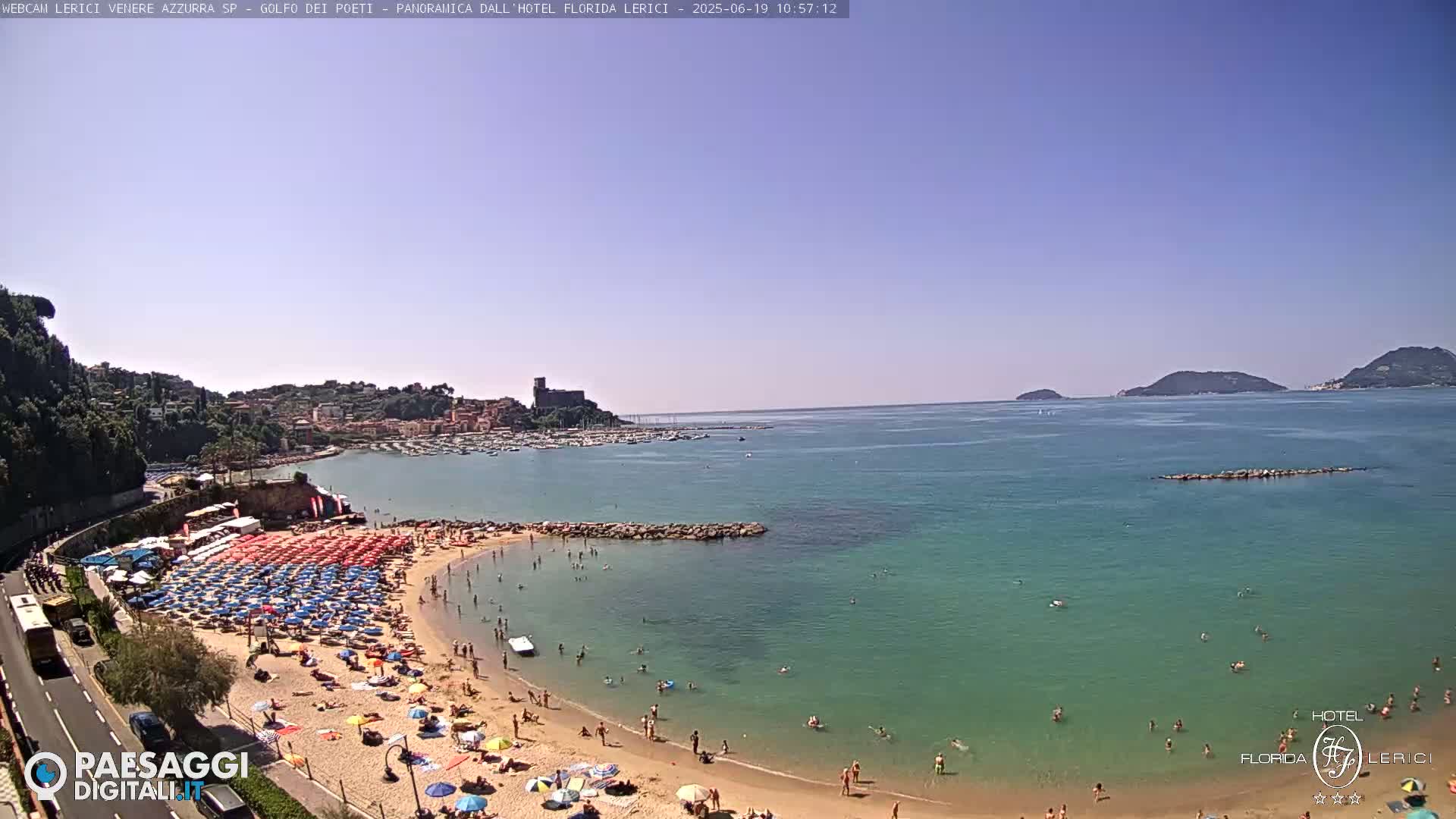 A sunny beach scene shows a calm, teal-colored ocean with a sandy cove crowded with people under colorful umbrellas, a town nestled into the hills beyond, and islands visible in the distance.