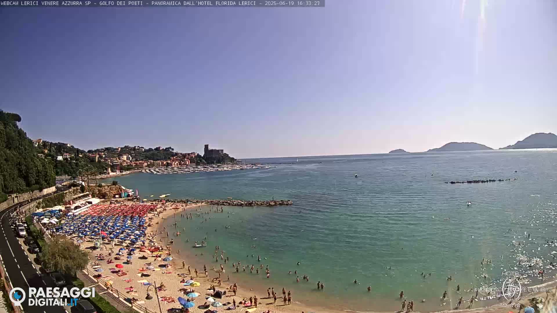A sunny beach scene shows a crowded sandy beach with many colorful umbrellas and people swimming in clear, teal water, with a town and distant hills visible in the background.