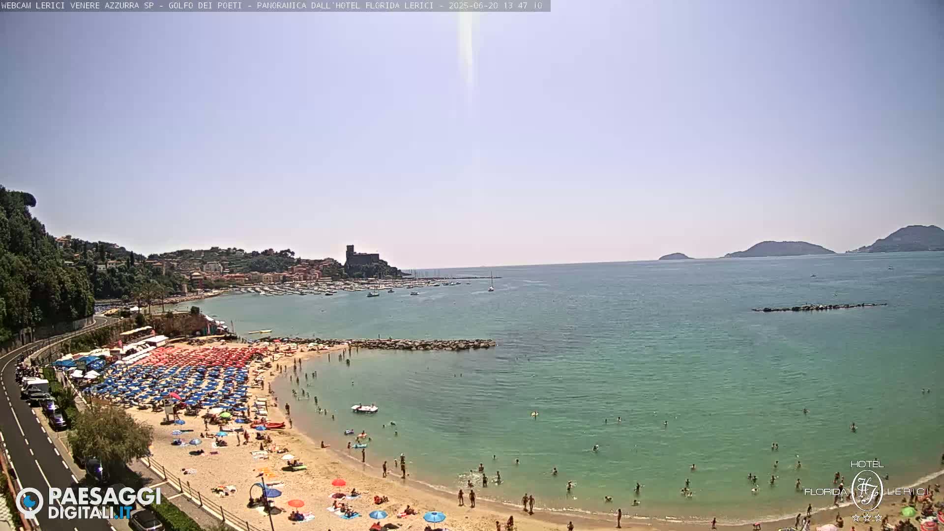 A sunny day at a crowded beach with clear, calm water, a small town, and boats in the bay, with distant hills in the background.