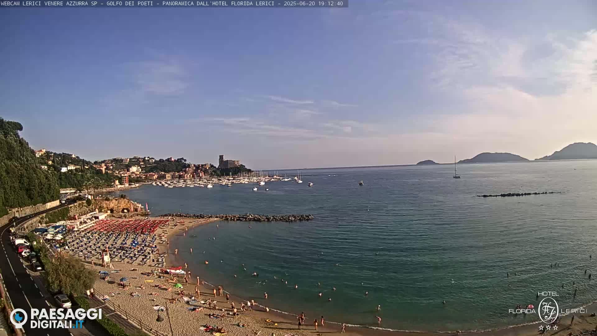 A sunny day at a sandy beach with clear water, many people sunbathing and swimming, boats in a harbor, and hills in the background.