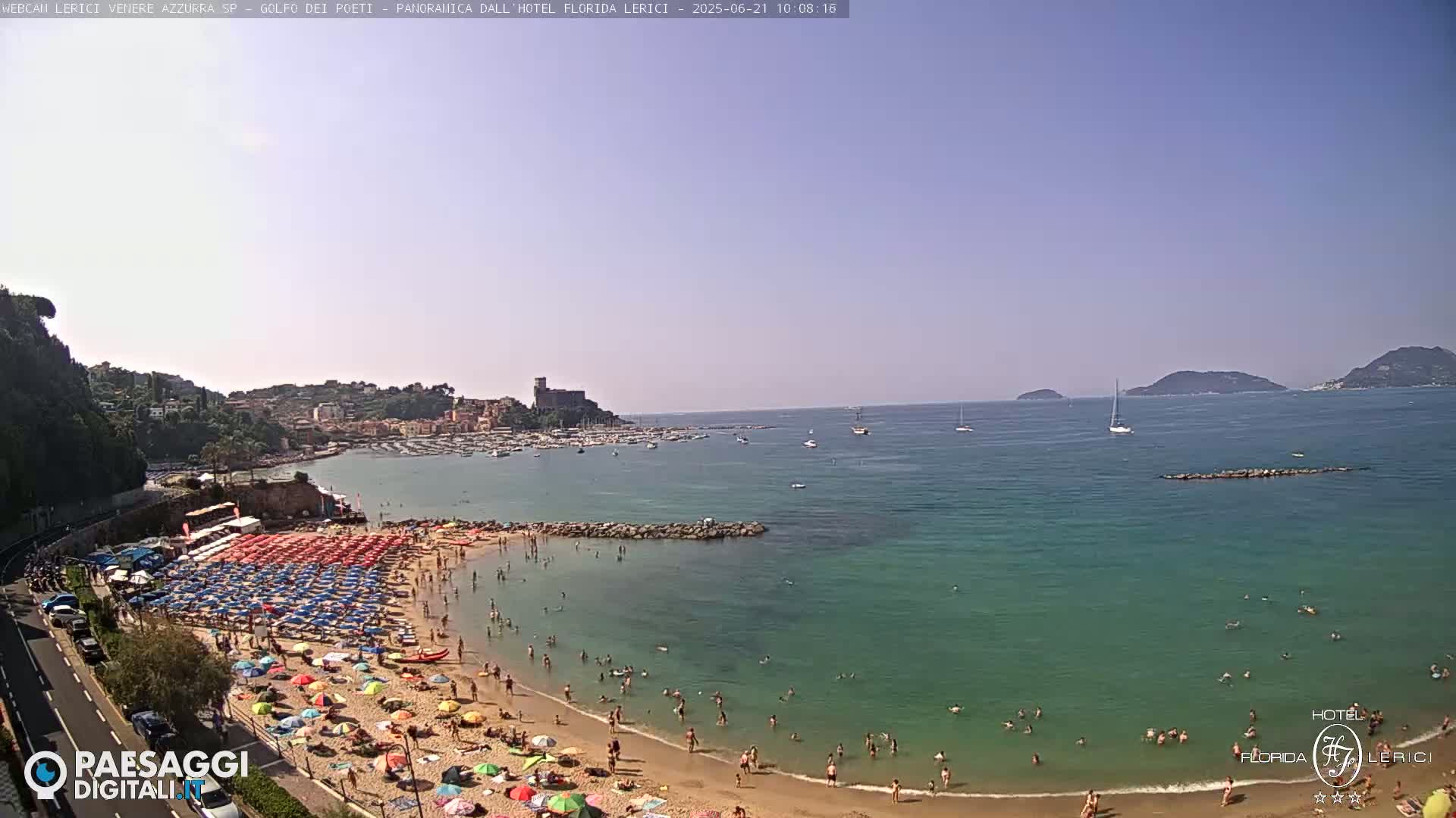 A sunny beach scene shows a sandy shore crowded with colorful umbrellas and bathers, calm turquoise water extending to a distant coastline with boats and islands.
