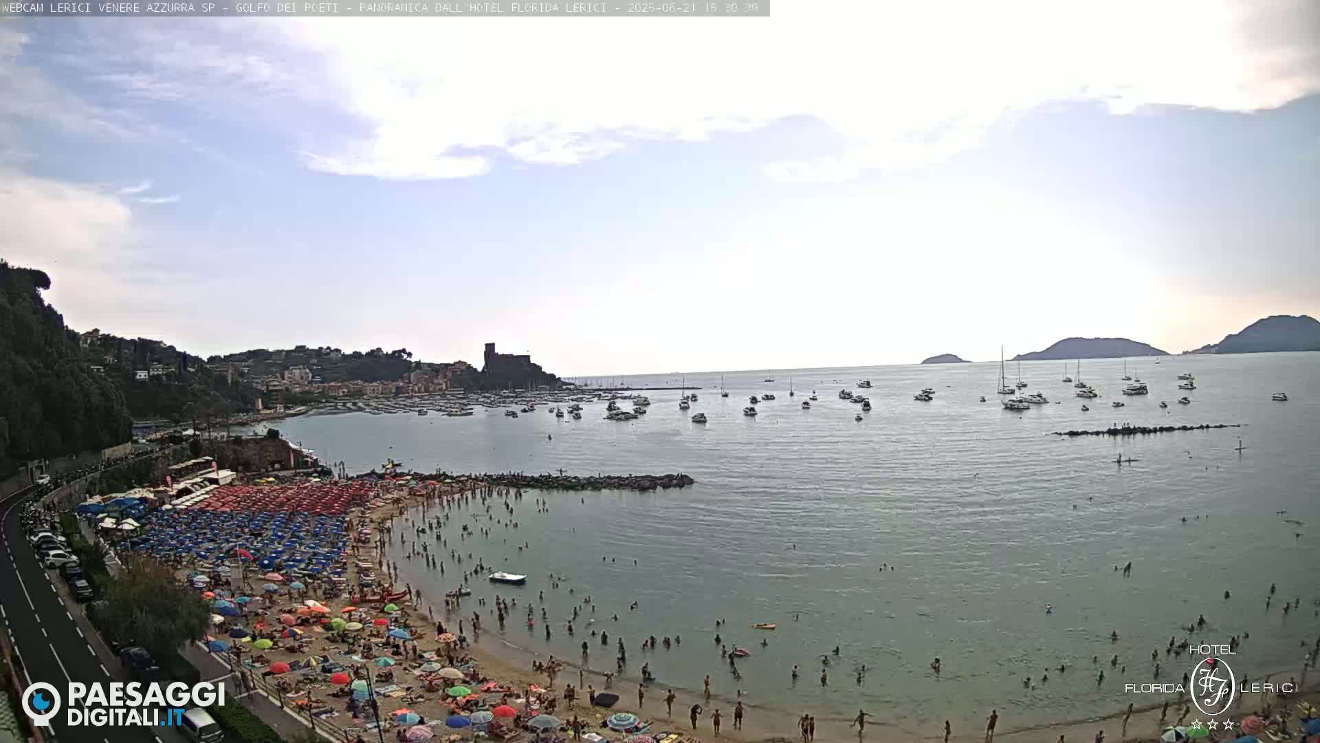 A crowded beach with numerous colorful umbrellas is situated next to a calm, light-green sea filled with boats under a partly cloudy sky.